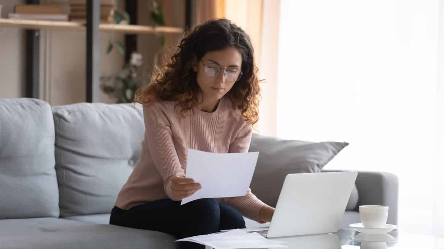 Focused young Caucasian woman in glasses sit on couch at desk work on laptop read paperwork, concentrated millennial businesswoman manage paper documents, pay bills or taxes, use computer at home