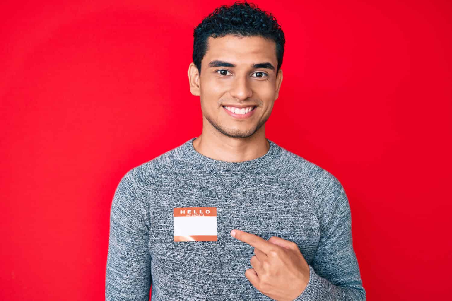Young handsome hispanic man wearing hello my name is sticker identification smiling happy pointing with hand and finger 
