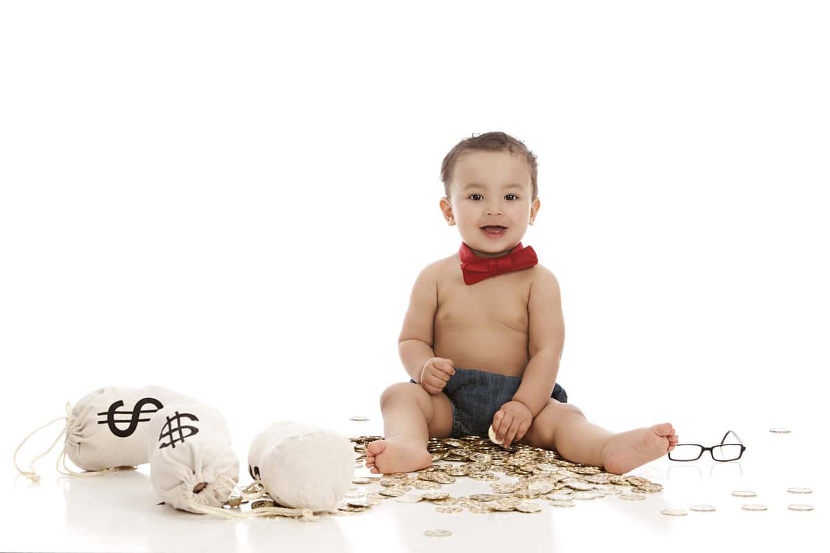 Future banker. Adorable baby boy wearing a red bow-tie and sitting on a pile of gold coins next to money bags. Isolated on white with room for your text.