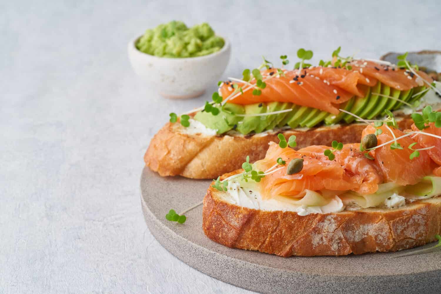 Two open sandwich, toast with salmon, cream cheese, avocado, cucumber slices on white concrete table. Morning healthy breakfast with fish and cup of drink, side view, copy space