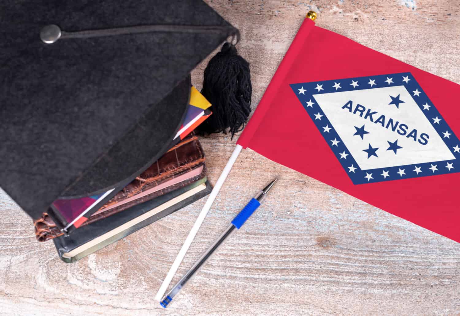 Black graduation hat on books next to Arkansas flag, education concept, top view