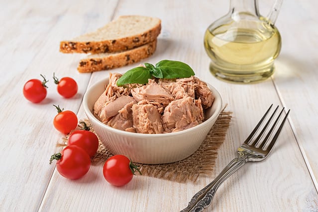 Canned tuna meat in a bowl, fork, bread and fresh red cherry tomatoes on a white wooden table. Low calories healthy eating snack of preserved tuna fish and vegetables. Tasty seafood. Front view.