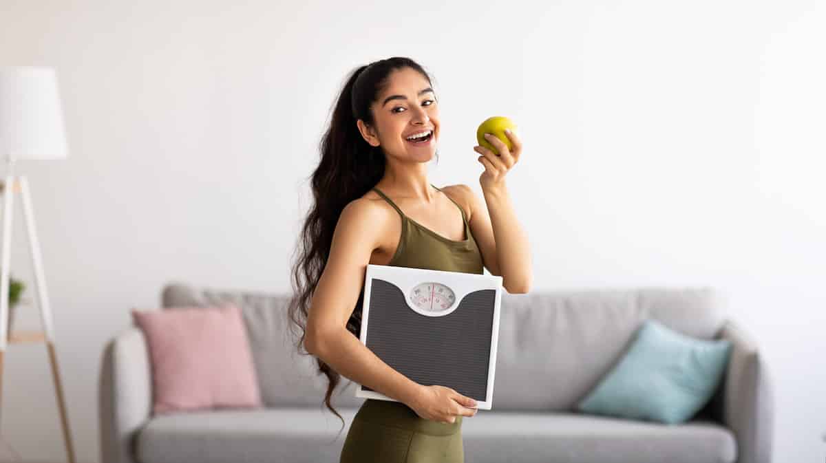 Portrait of millennial Indian lady holding scales and eating apple, choosing healthy diet, panorama. Young Asian woman preferring wholesome nutrition. Wellbeing and weight loss concept