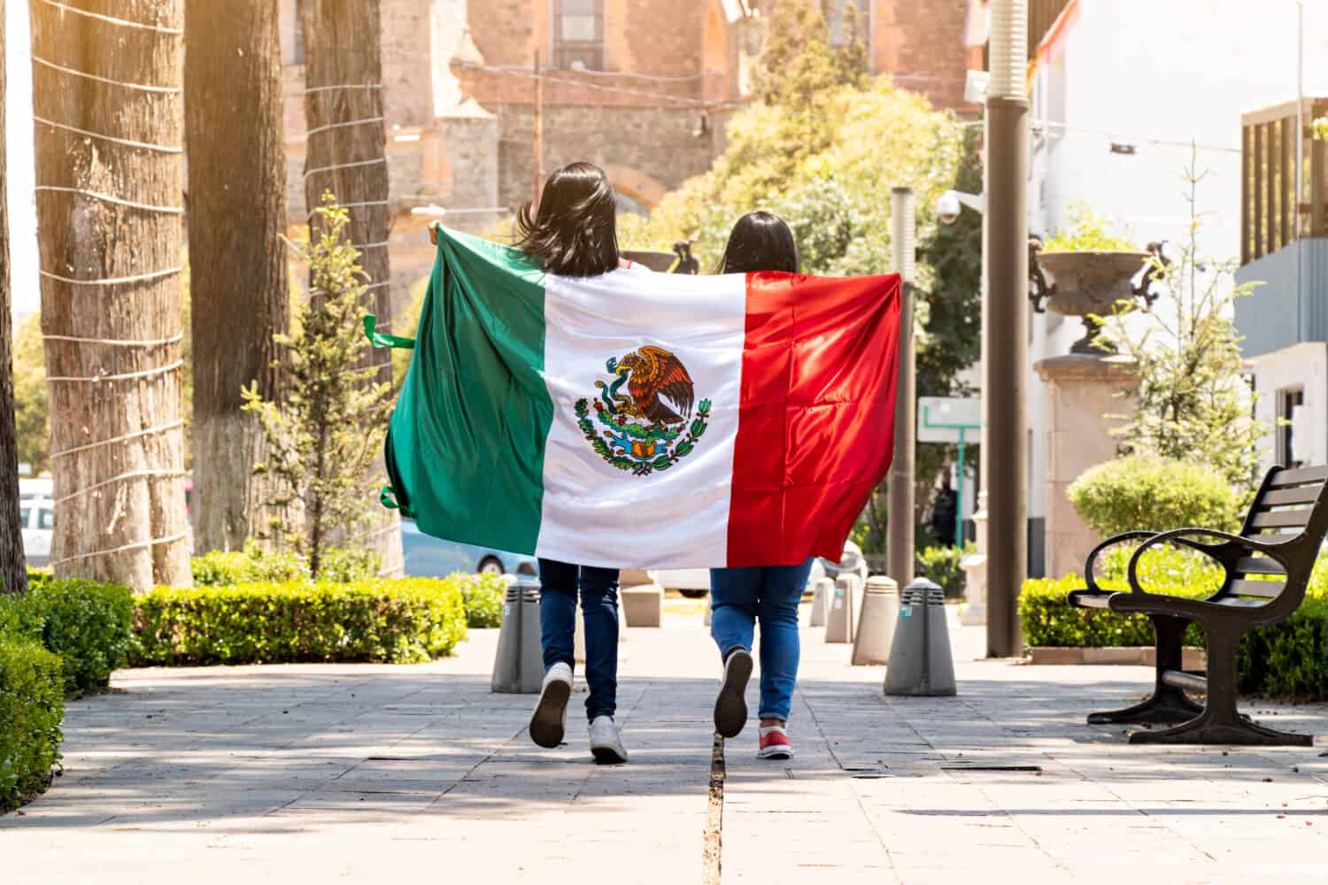 two young mexican women running on a walkway on a tree-lined street, with the mexican flag