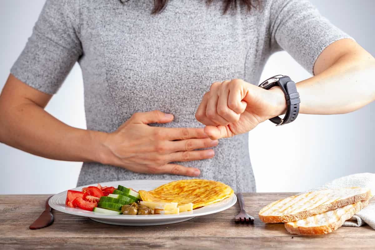 Intermittent fasting concept with a woman sitting hungry in front of food and looking at her watch to make sure she breaks fast on the correct time. A dietary modification for healthy lifestyle.