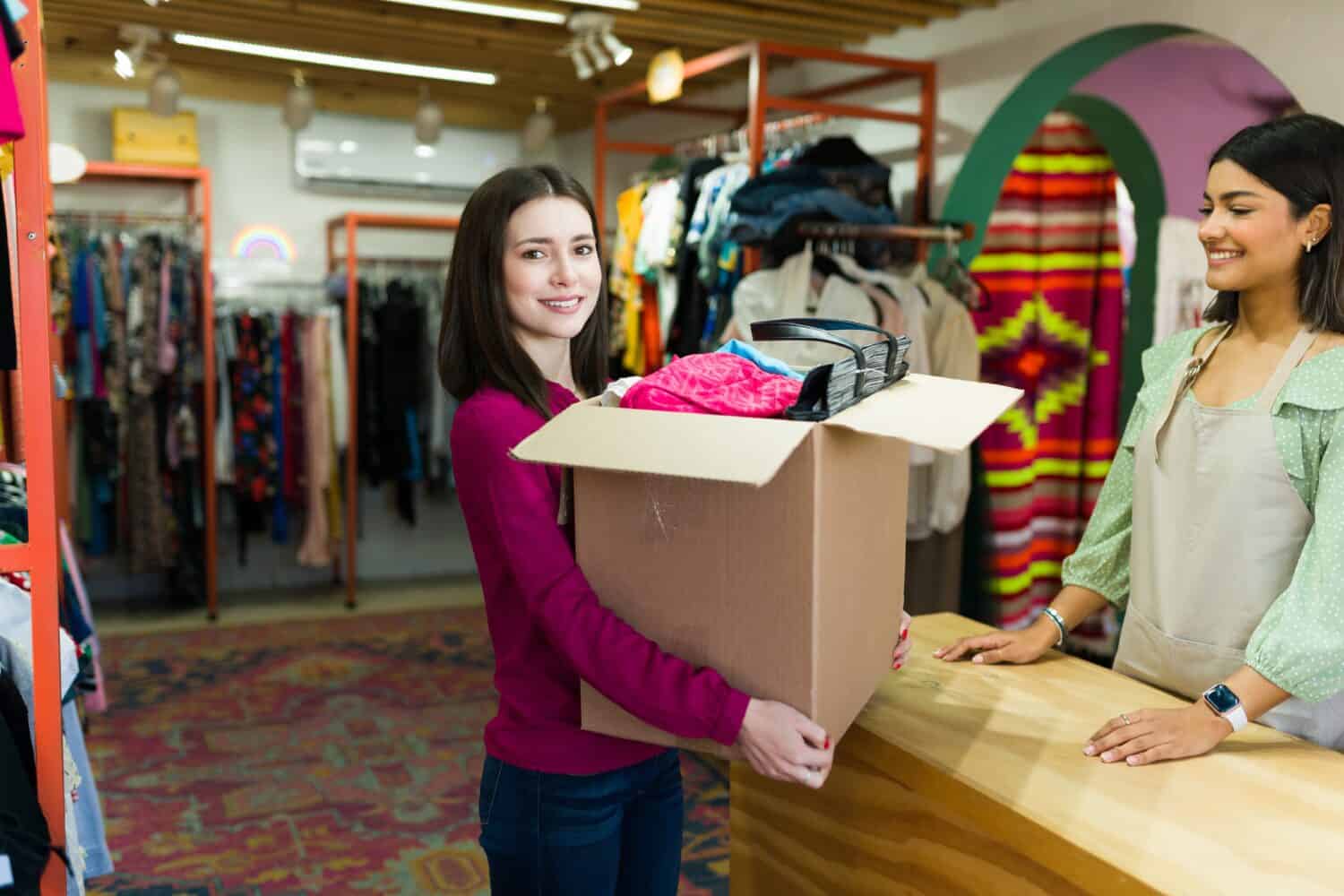 Beautiful young woman carrying a box while selling her clothes and bags at the thrift store. Smiling woman selling second hand clothing