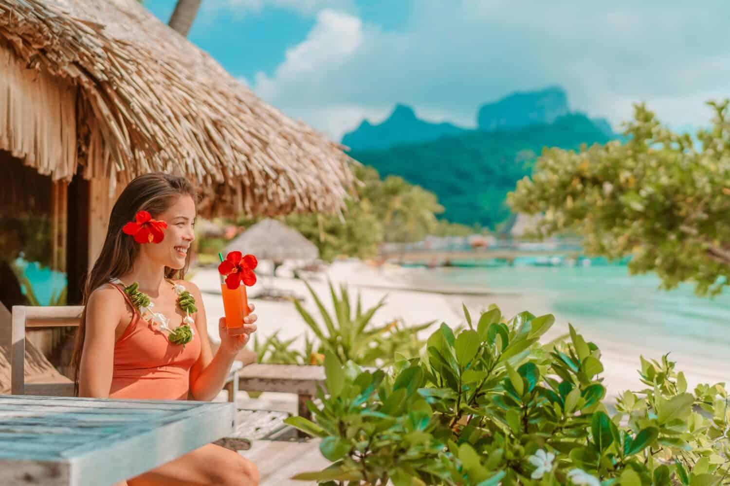 Breakfast at luxury hotel room on beach. Asian woman drinking fruit juice morning on summer vacation travel in Bora Bora island, Tahiti French Polynesia landscape. Happy tourist relaxing on holiday.