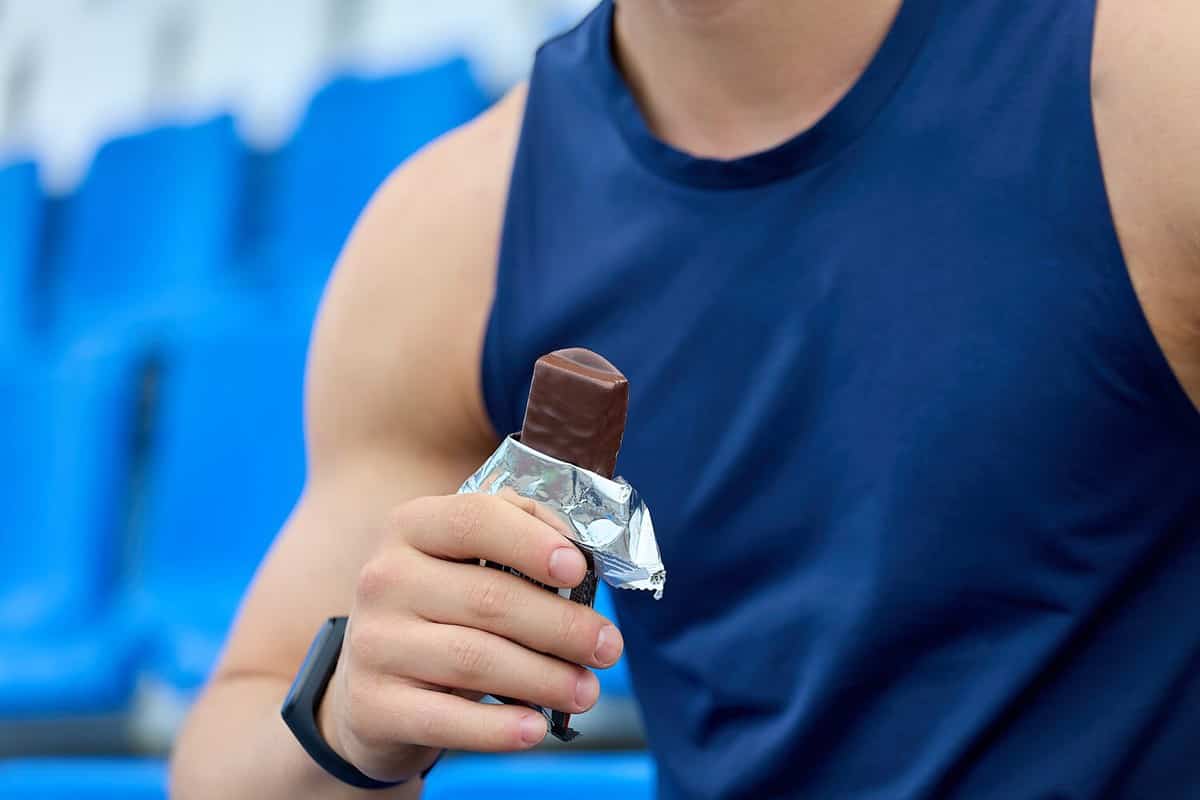 Muscular athlete snacks on a protein bar while resting after a workout at the stadium. Man in a blue sports jersey rests in the stand for fans, eating a chocolate bar.
