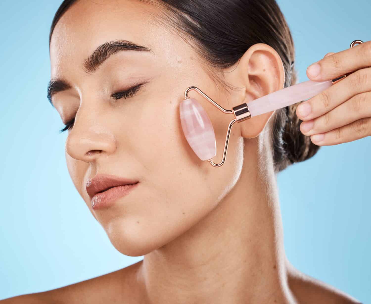 Skin care, face roller and beauty woman with facial massage for dermatology, cosmetic and wellness. Young aesthetic model person for natural rose quartz spa product to relax on blue background