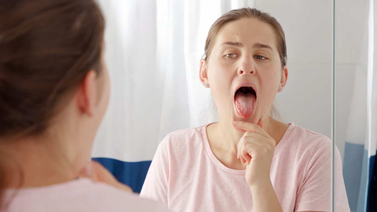 Portrait of young brunette woman checking her tongue for plaque and microbes while looking in mirror.