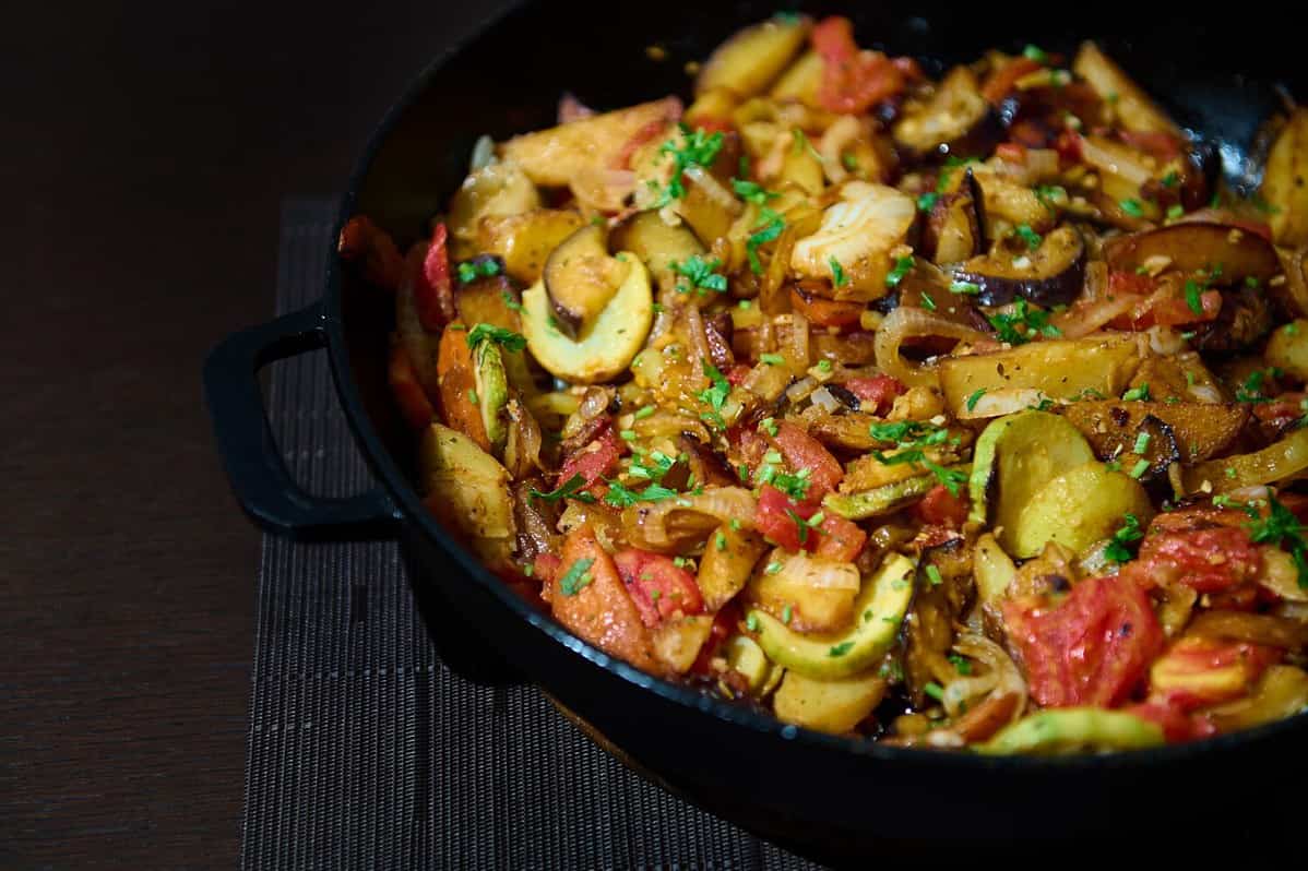 Freshly cooked vegetable stew of seasonal organic veggies sprinkled with fresh herbs, in a cast-iron skillet, on wooden background. Vegetarian meal. Healthy eating and dieting concept. Food still life