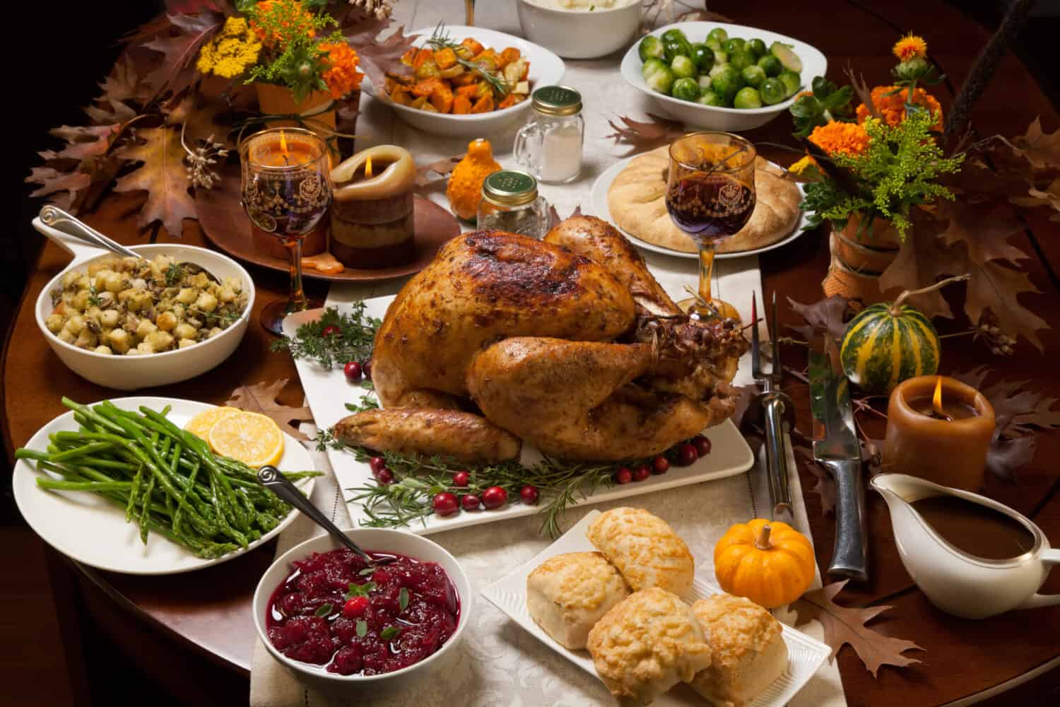 Roasted turkey garnished with cranberries on a rustic style table decoraded with pumpkins, gourds, asparagus, brussel sprouts, baked vegetables, pie, flowers, and candles.