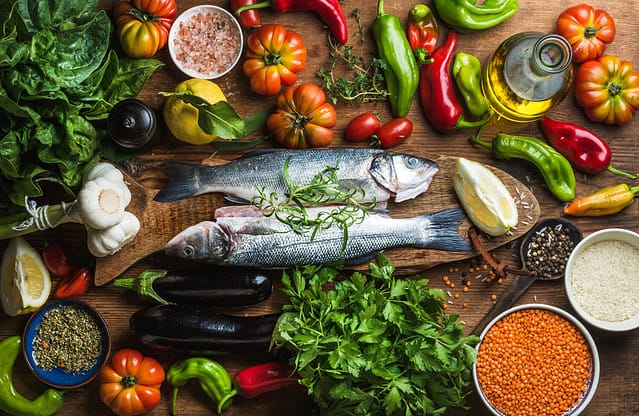 Raw uncooked seabass fish with vegetables, grains, herbs and spices on chopping board over rustic wooden background, top view