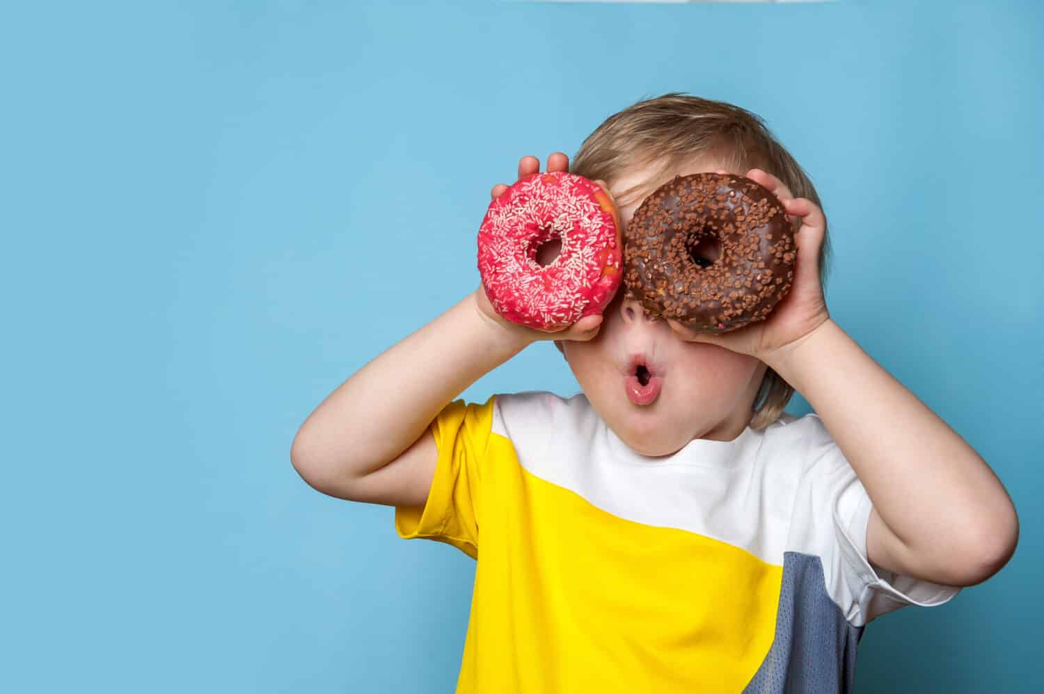 Little happy cute boy is eating donut on blue background wall. child is having fun with donut. Tasty food for kids. Funny time at home with sweet food. Bright kid.