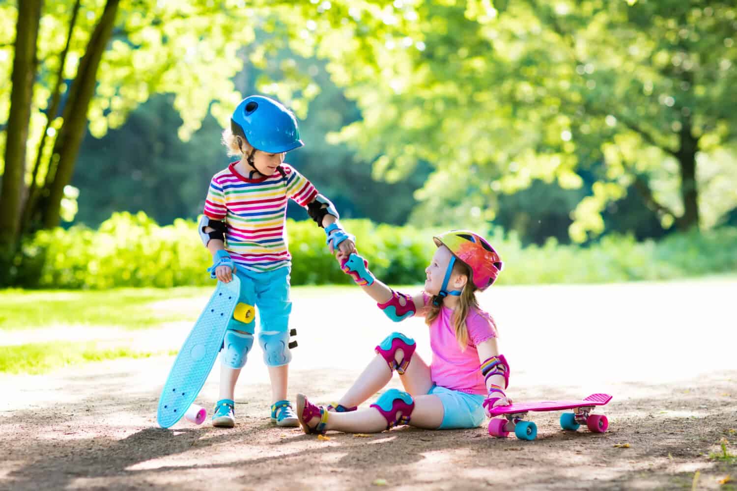 Children riding skateboard in summer park. Little girl and boy learn to ride skate board, help and support each other. Active outdoor sport for kids. Child skateboarding. Preschooler kid skating.