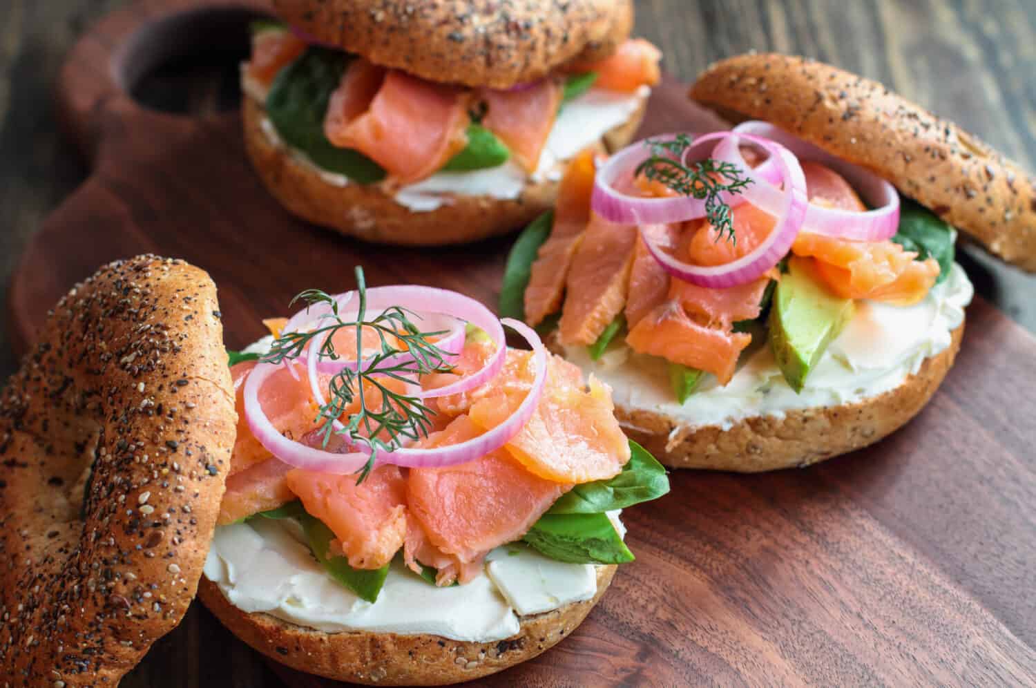 Lox - Everything bagel with smoked salmon, spinach, red onions, avocado and cream cheese over a rustic wood table background. Selective focus with blurred background.