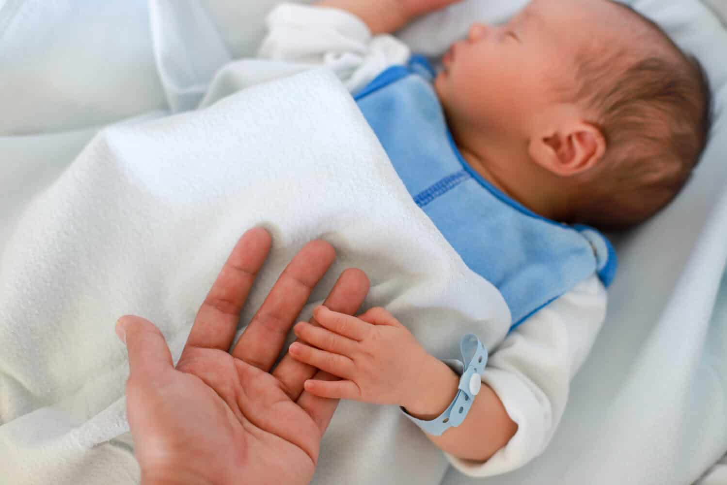 Mother holding her newborn baby hand with name tag bracelets. infant boy first day of life in bassinet bed at hospital.