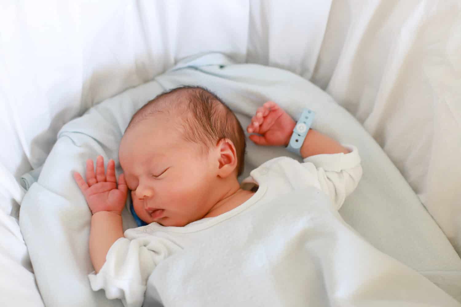 Newborn baby with name tag bracelets sleeping in bassinet bed at hospital. Mixed race Asian-German infant first day of life.