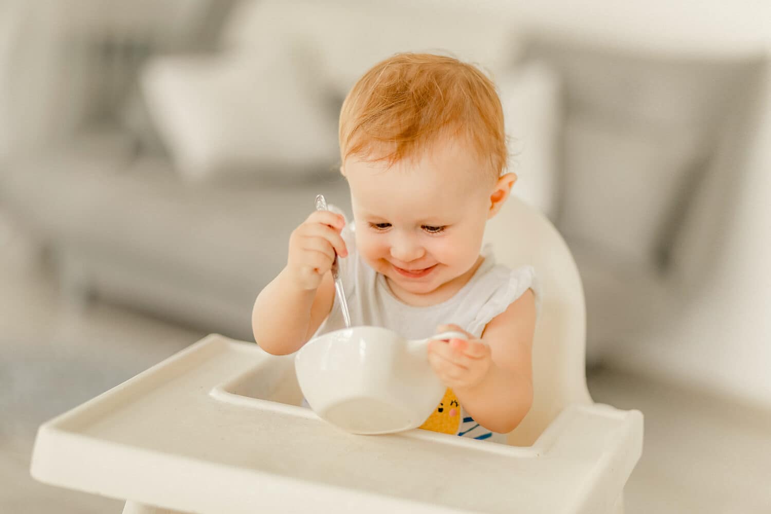 A little girl under the age of 1 year in a bright kitchen in a white highchair sits and eats from a white plate.