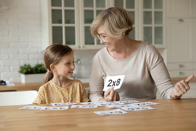 Smiling mature grandmother with little granddaughter learning multiplication table using flash cards sitting at kitchen table, middle aged female teacher helping to cute kid schoolgirl with math