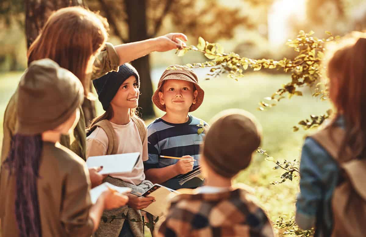Group of curious school children with notebooks listening to their young female teacher while learning about nature together, looking at green leaf during ecology lesson in autumn forest on sunny day