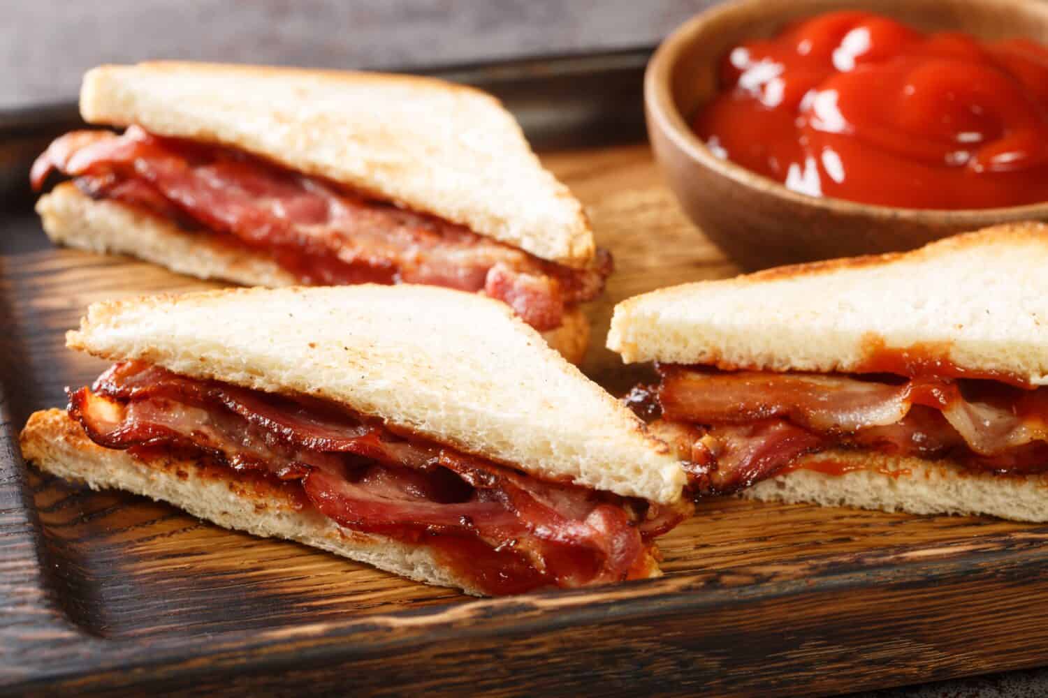 Bacon butty is a British sandwich consisting of crispy bacon, butter, and sauce closeup in the wooden tray on the table. Horizontal