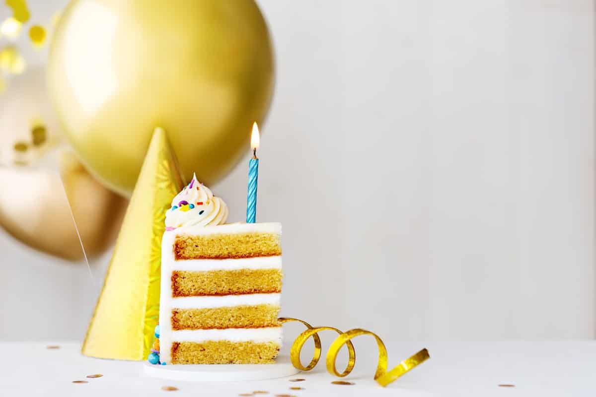 Slice of birthday cake with single blue birthday candle and gold birthday balloons