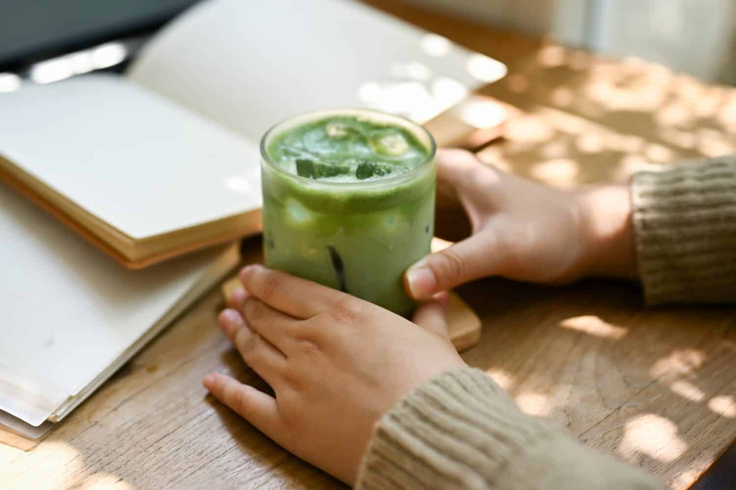 Close-up image of a female in comfy sweater holding a glass of iced matcha green tea at a table in a beautiful minimal cafe.