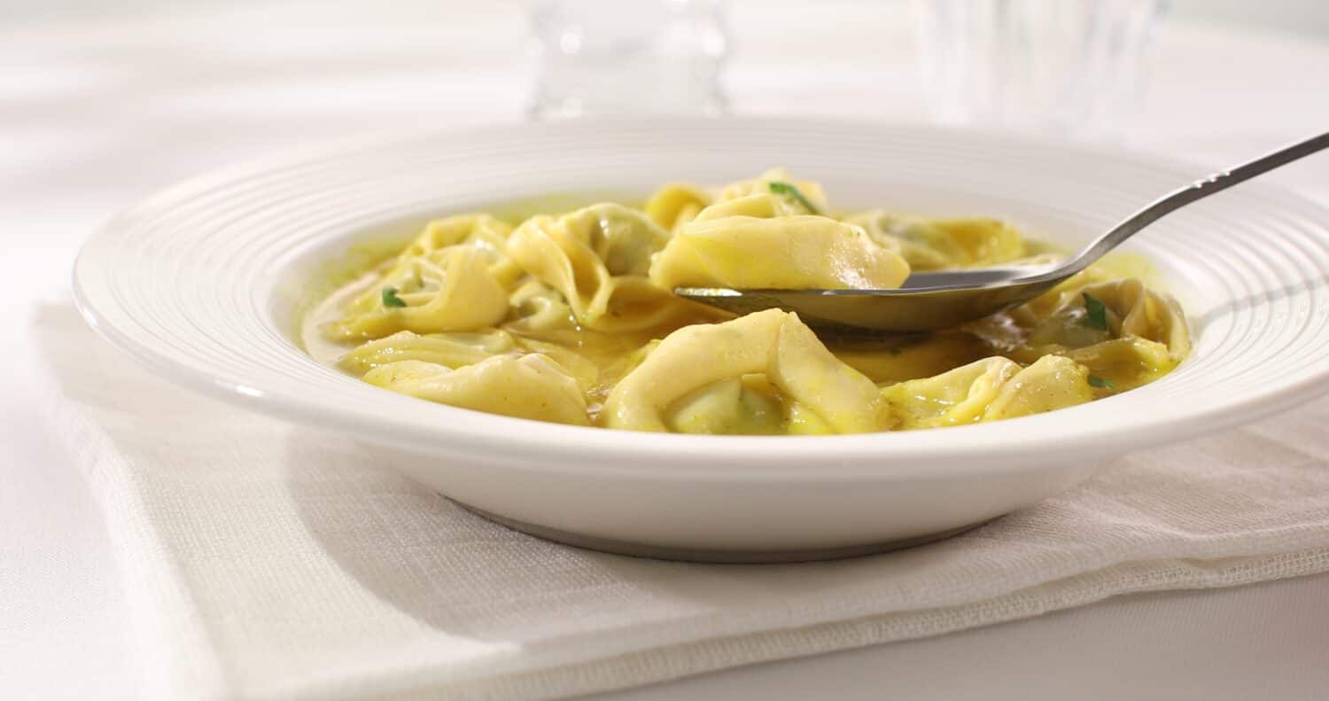 spoon takes tortelloni with ricotta and spinach in chicken broth. italian pasta on a white plate. ready to eat food. mediterranean cuisine. sunny day. angle view.