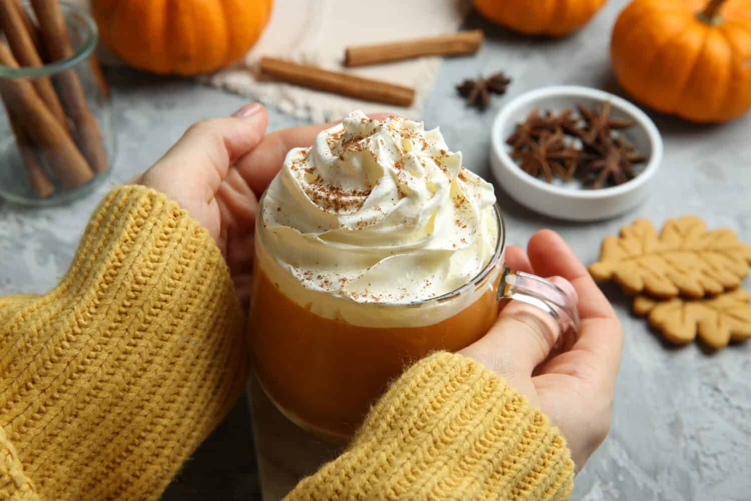 Woman holding cup of pumpkin spice latte with whipped cream at light grey table, closeup