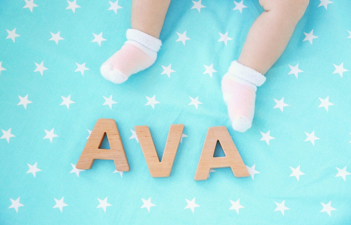Legs of cute baby and word AVA composed of wooden letters on bed. Choosing name concept