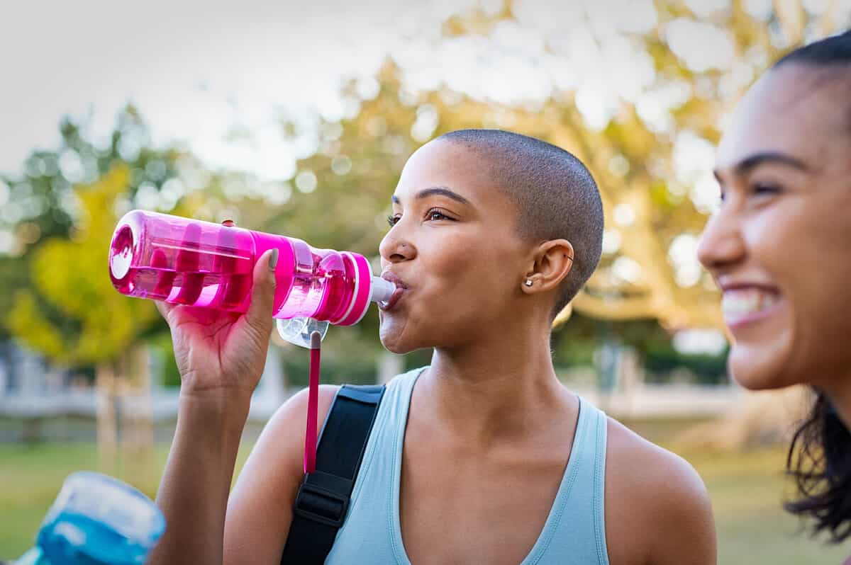 Sporty woman drinking water from bottle after fitness exercise. Young bald woman sipping water from pink bottle after workout at park with friends. Closeup face of african american girl refreshing.