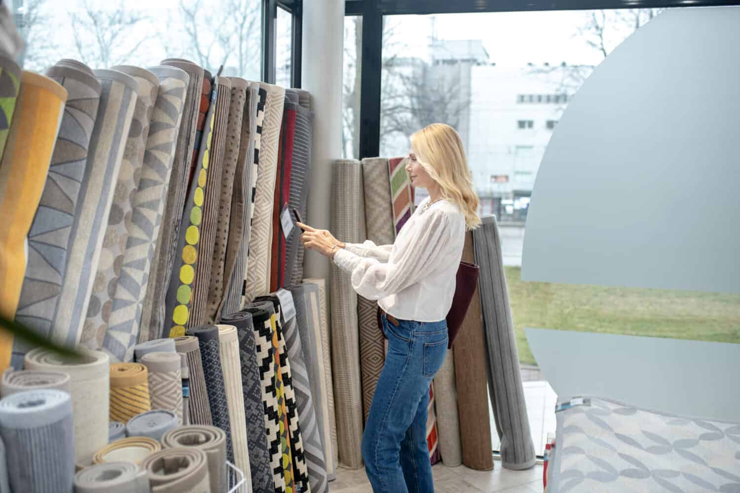 Carpets, a choice. Woman with hair to the shoulders with a smartphone standing in front of the carpets and taking a photo in the furniture salon.