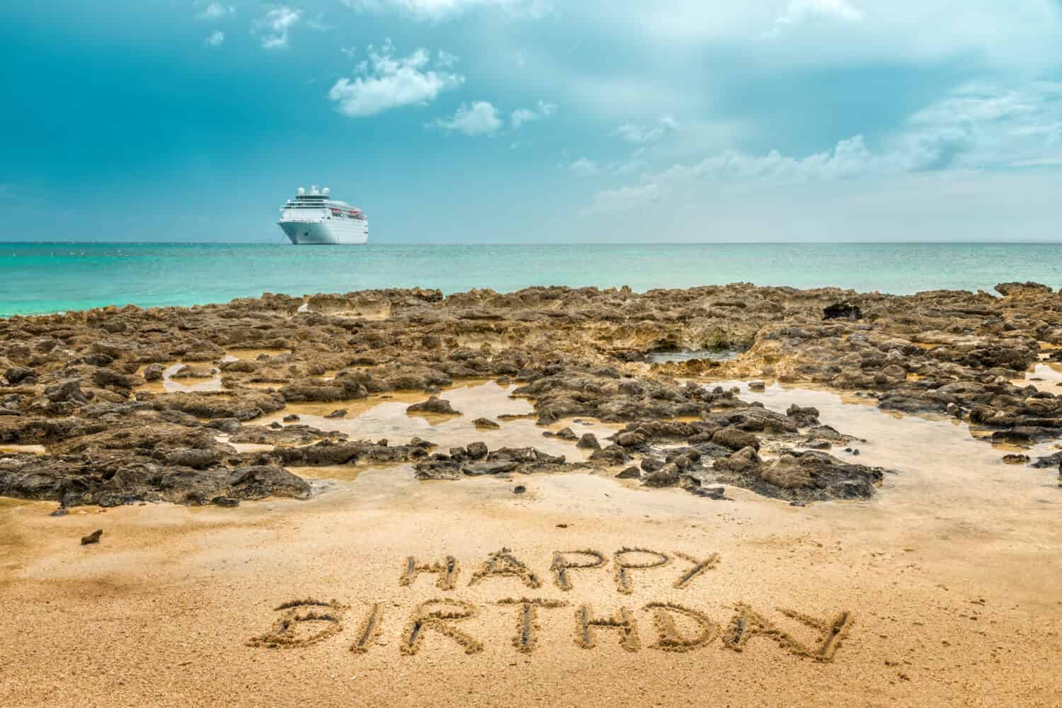 Hand written  "Happy Birthday" on the sandy beach by the ocean with a cruise ship in the background