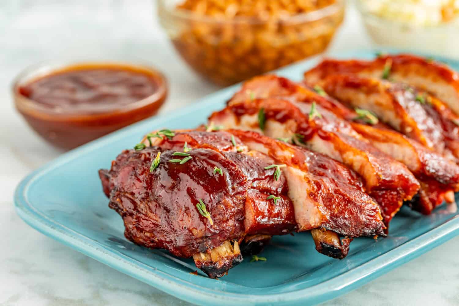 Close up of rack of bbq smoked ribs sliced on bright blue plate with sides of cole slaw and backed beans in background and bowl of sauce