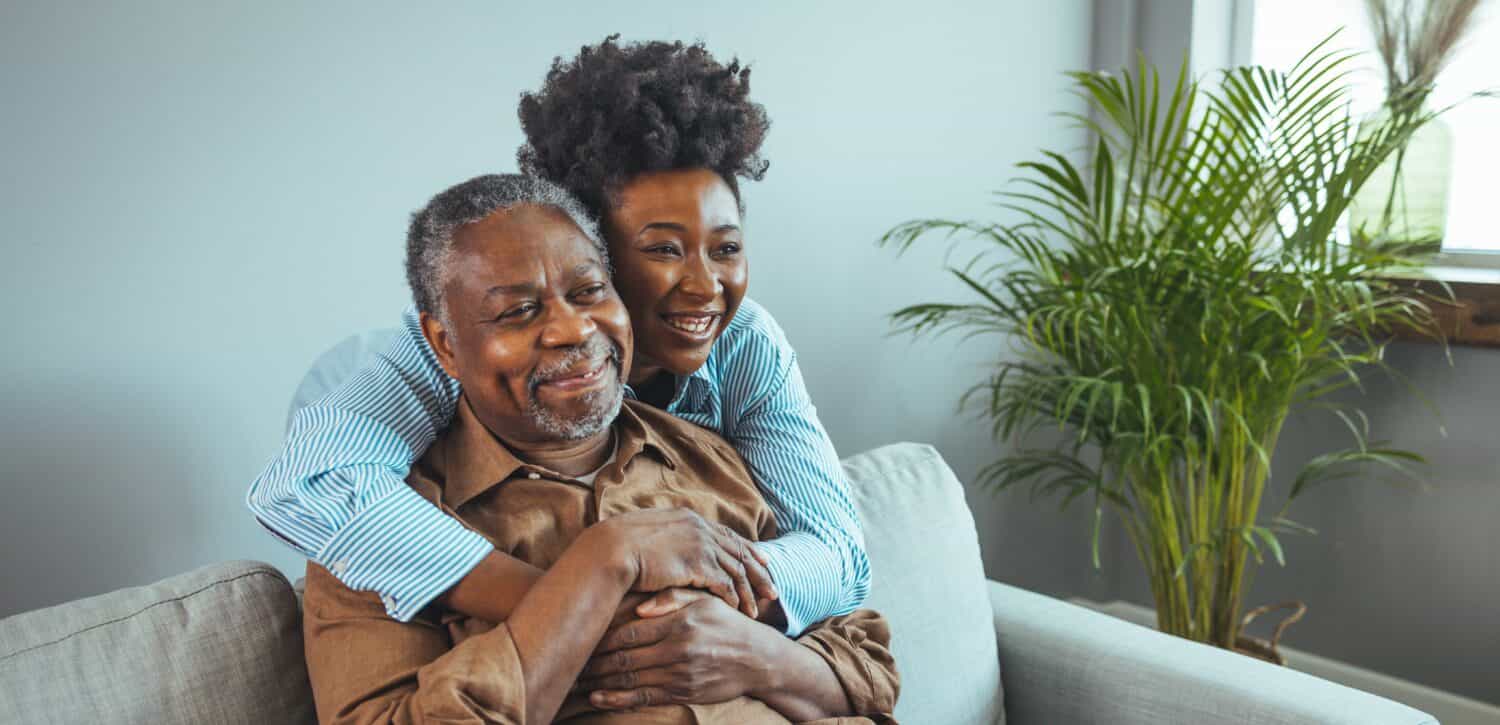 Adult daughter visits senior father in assisted living home. Portrait of a daughter holding her elderly father, sitting on a bed by a window in her father's room. 