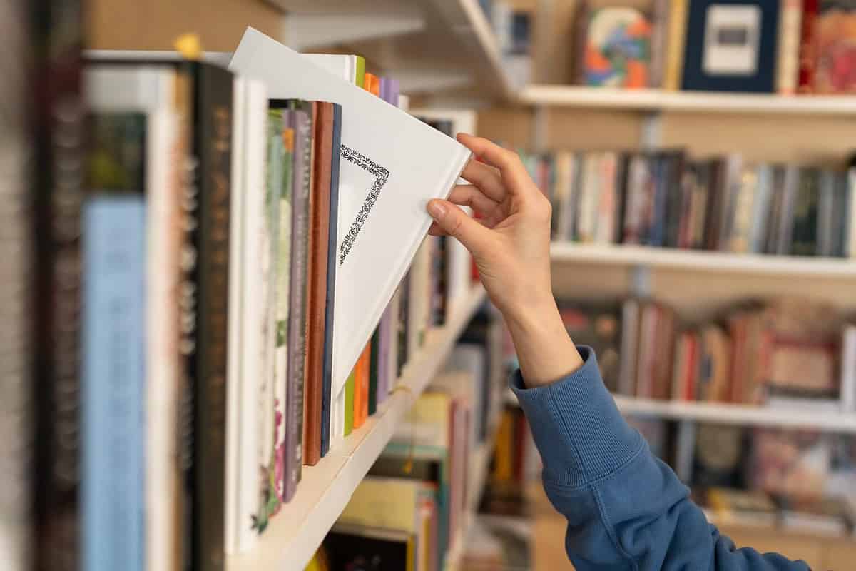 Female hand pulling book from bookshelf in public library in university, college or high school. Woman student take novel from bookcase in bookshop store, soft focus. Education and literature concept