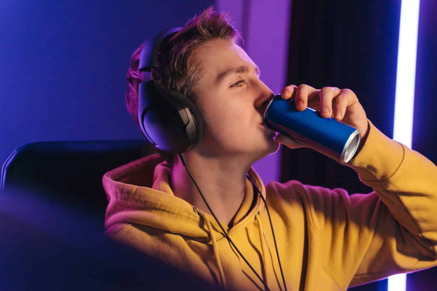 Young professional cybersport gamer playing online video game late at night, drinking caffeine energy drink to wake up and concentrate, focus on multiplay tournament. Games addiction concept