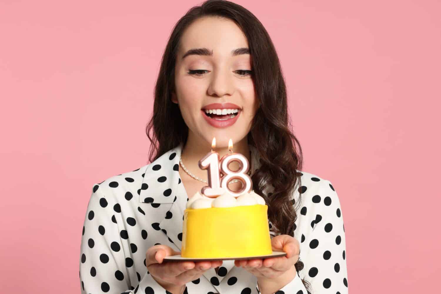 Coming of age party - 18th birthday. Smiling woman holding delicious cake with number shaped candles on pink background