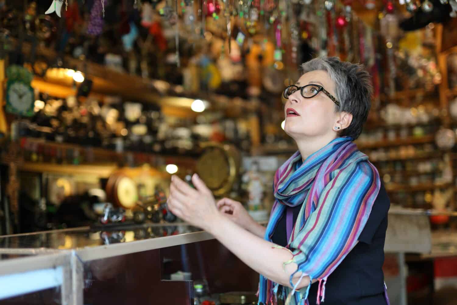 A cheerful mature woman browses for antique items at a flea market store. She is wearing a colorful scarf and looking at the display case with various jewelry.