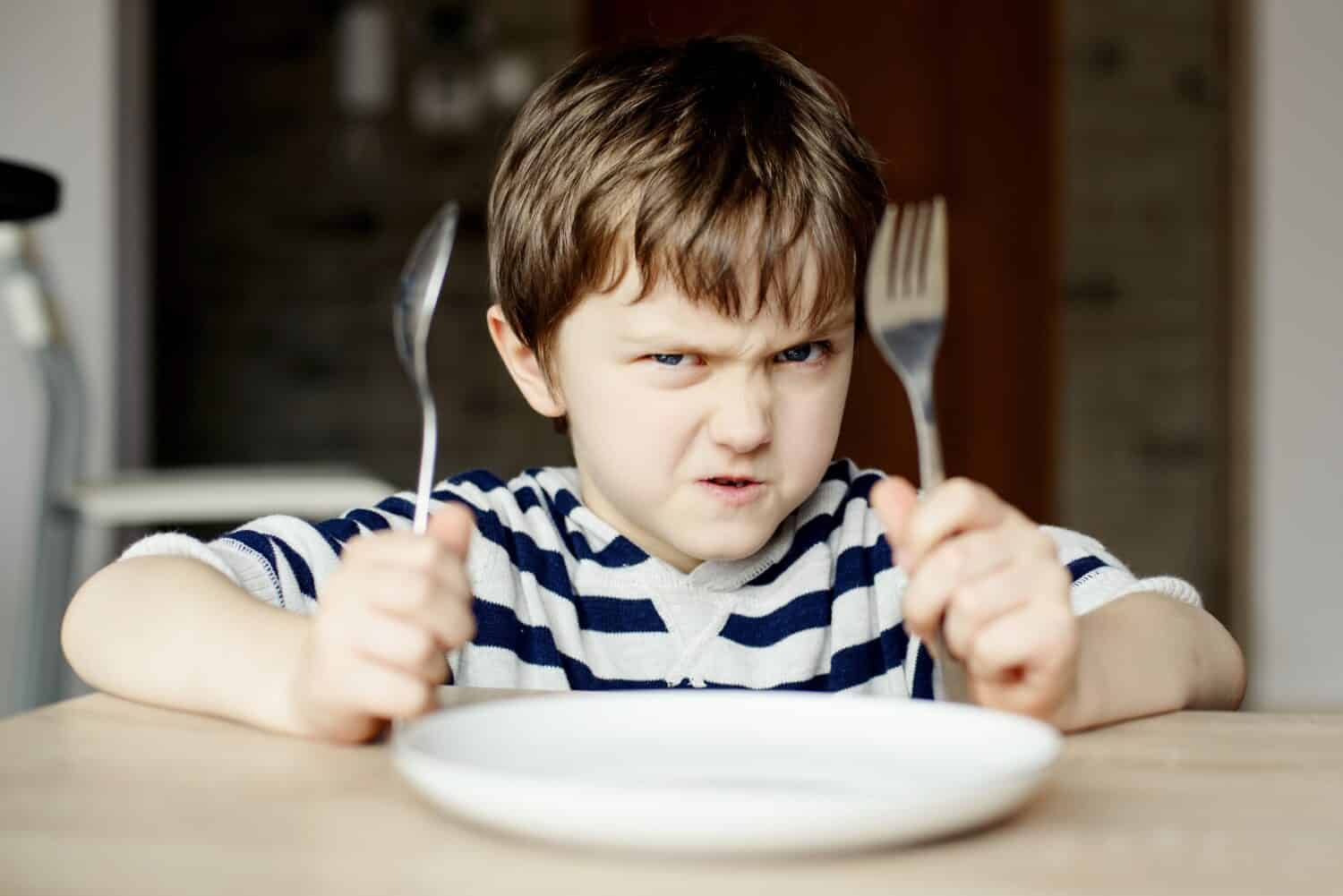 Furious little boy waiting for dinner. Holding a spoon and fork in the hand