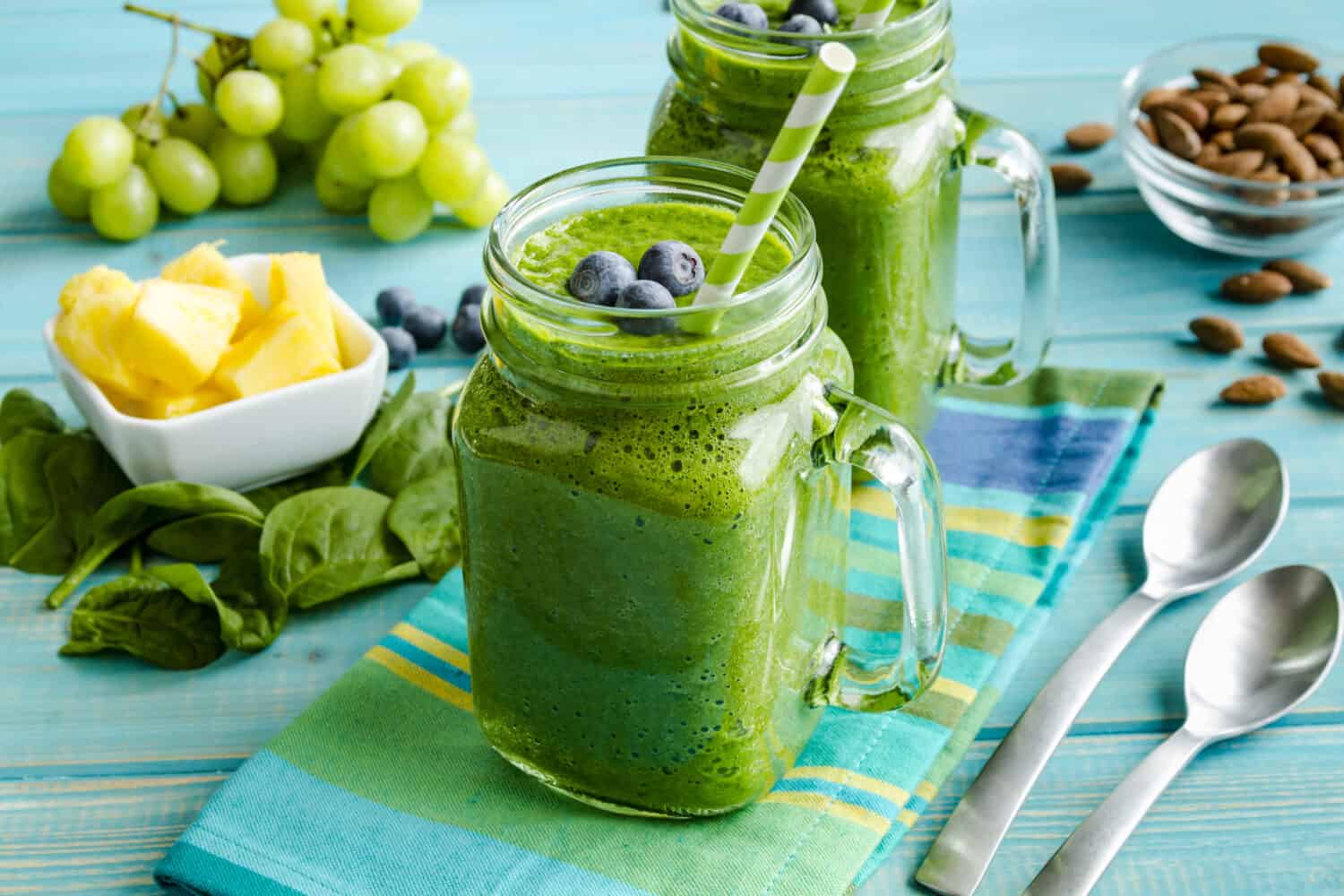 Mason jar mugs filled with green spinach and kale health smoothie with green swirled straw sitting with blue striped napkin and spoons