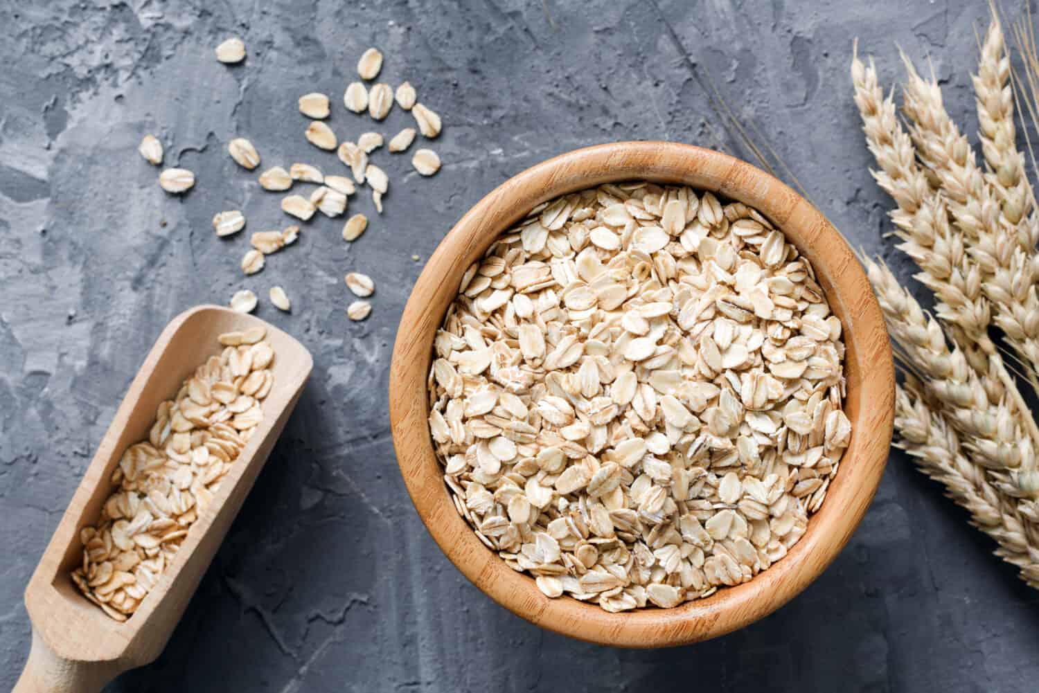 Rolled oats or oat flakes in wooden bowl and golden wheat ears on stone background. Top view, horizontal. Healthy lifestyle, healthy eating, vegan food concept