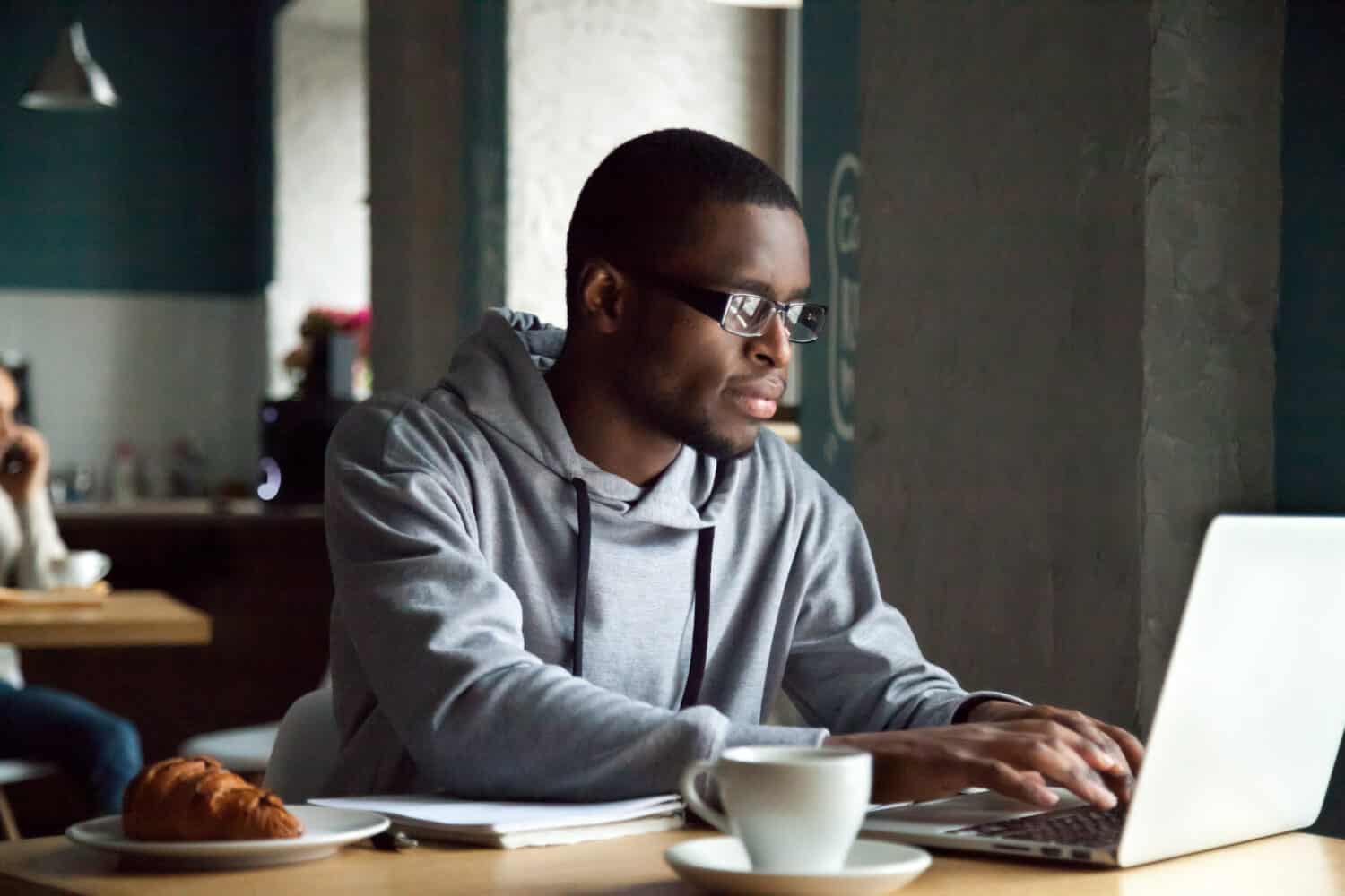 Serious millennial african-american man using laptop sitting at cafe table, focused black casual guy communicating online, writing emails, distantly working or studying on computer in public place