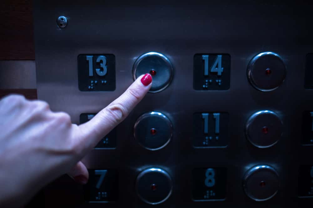 The hand of a woman with red fingernails presses the Number 13 on the elevator. Many elevators leave this number out due to beliefs that it is unlucky.