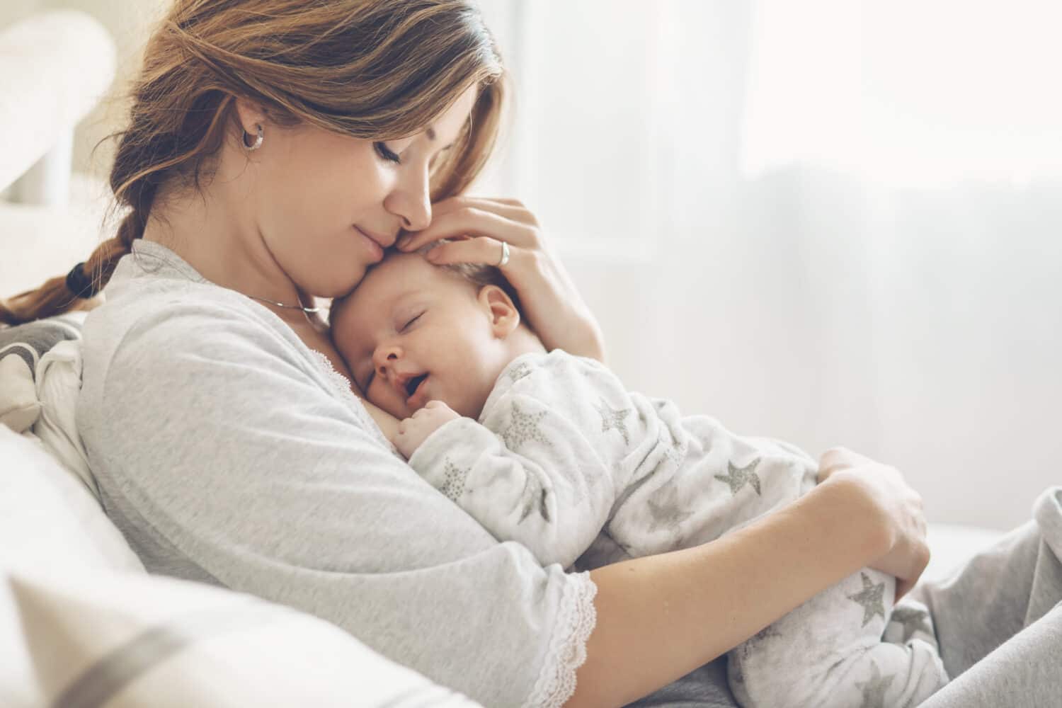 Loving mom carying of her newborn baby at home. Bright portrait of happy mum holding sleeping infant child on hands. Mother hugging her little 2 months old son.