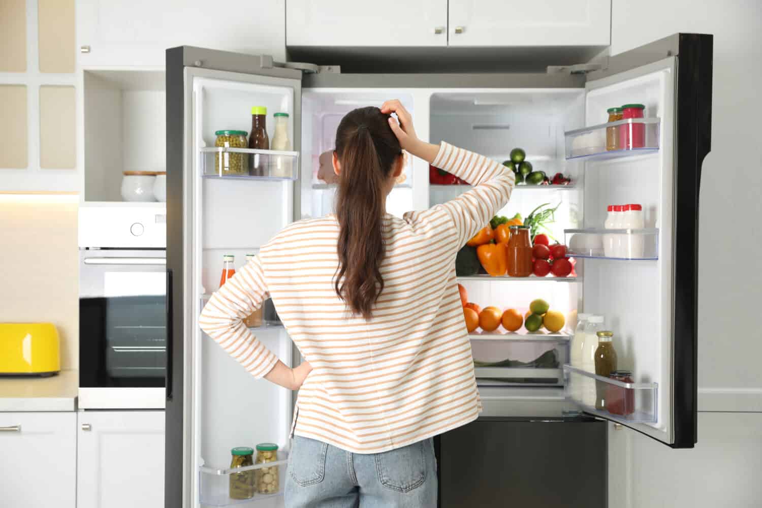 Young woman near open refrigerator in kitchen, back view