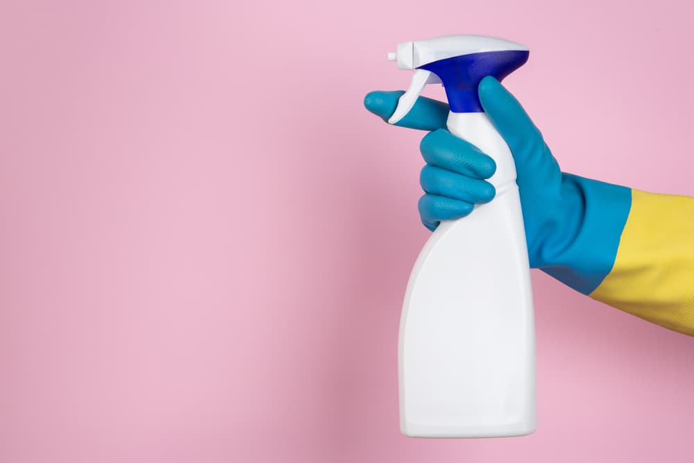 A hand with cleaning gloves squeezing a spray bottle on a pink background