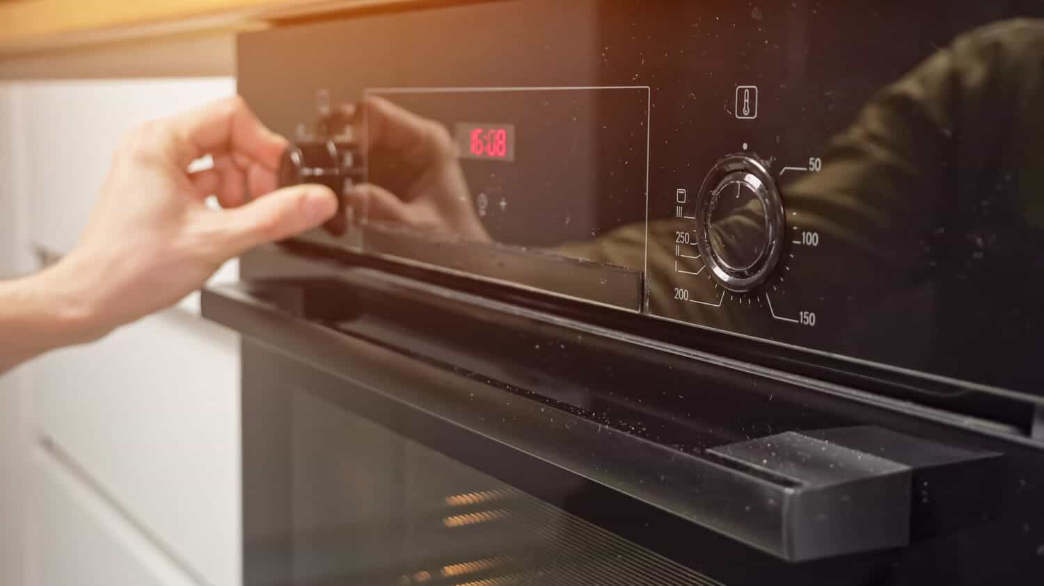 Man adjusts temperature and sets operating mode on modern oven in kitchen. Housekeeper prepares dinner using cooking appliance at home, sunlight