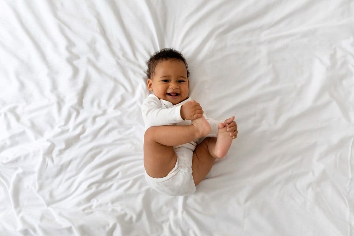 Cute Funny Little Black Baby Having Fun While Lying On Bed At Home, Adorable African American Infant Boy Playing With His Toes And Laughing While Resting In Bedroom, Top View, Copy Space